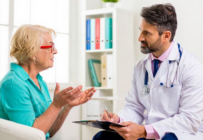Пожилая женщина в бирюзовой блузке и красных очках активно жестикулирует, рассказывая о симптомах врачу